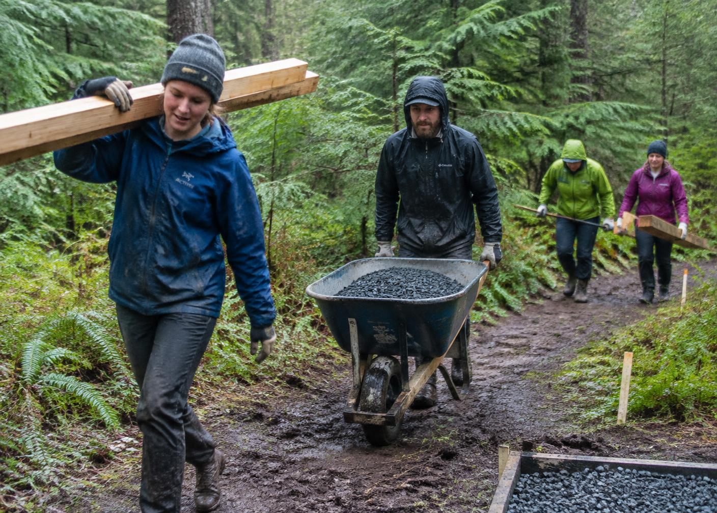 Volunteers in rain jackets carrying lumber and a wheelbarrow of gravel to build a tee pad